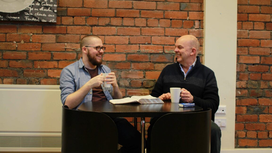 Two males chatting and smiling in a cafe holding cups of coffee with a book open.