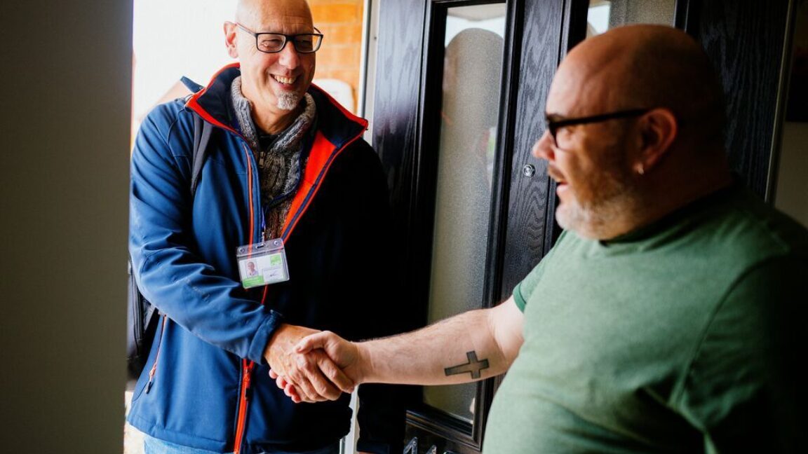 a man in a blue coat standing in a doorway, shaking hands with the man inside the house