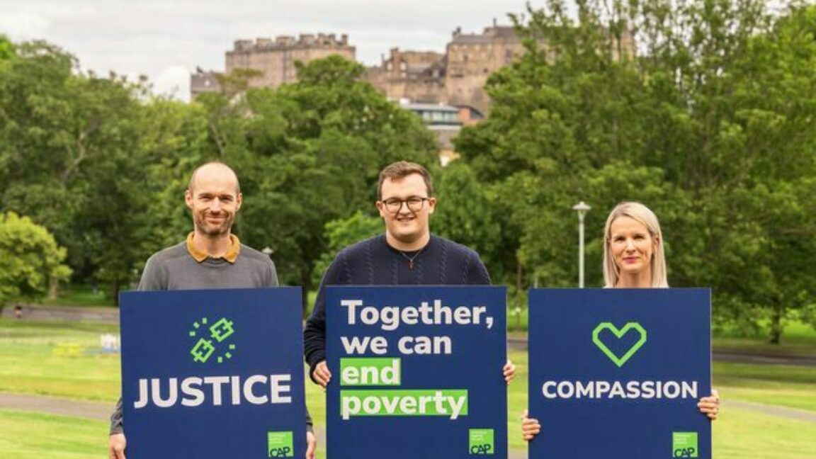 Two men and a woman, standing in a park with a Scottish castle behind them. Each holds a sign. The first reads justice, the second reads together we can end UK poverty, and the third reads compassion.