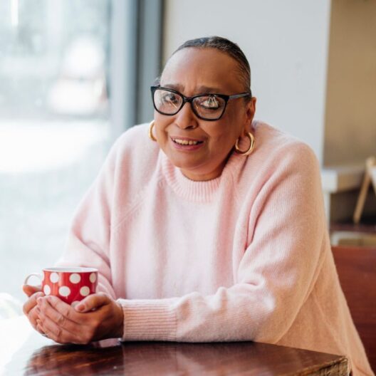 CAP client, Sharon, sat at a table holding a red mug, wearing a pink jumper and smiling at the camera