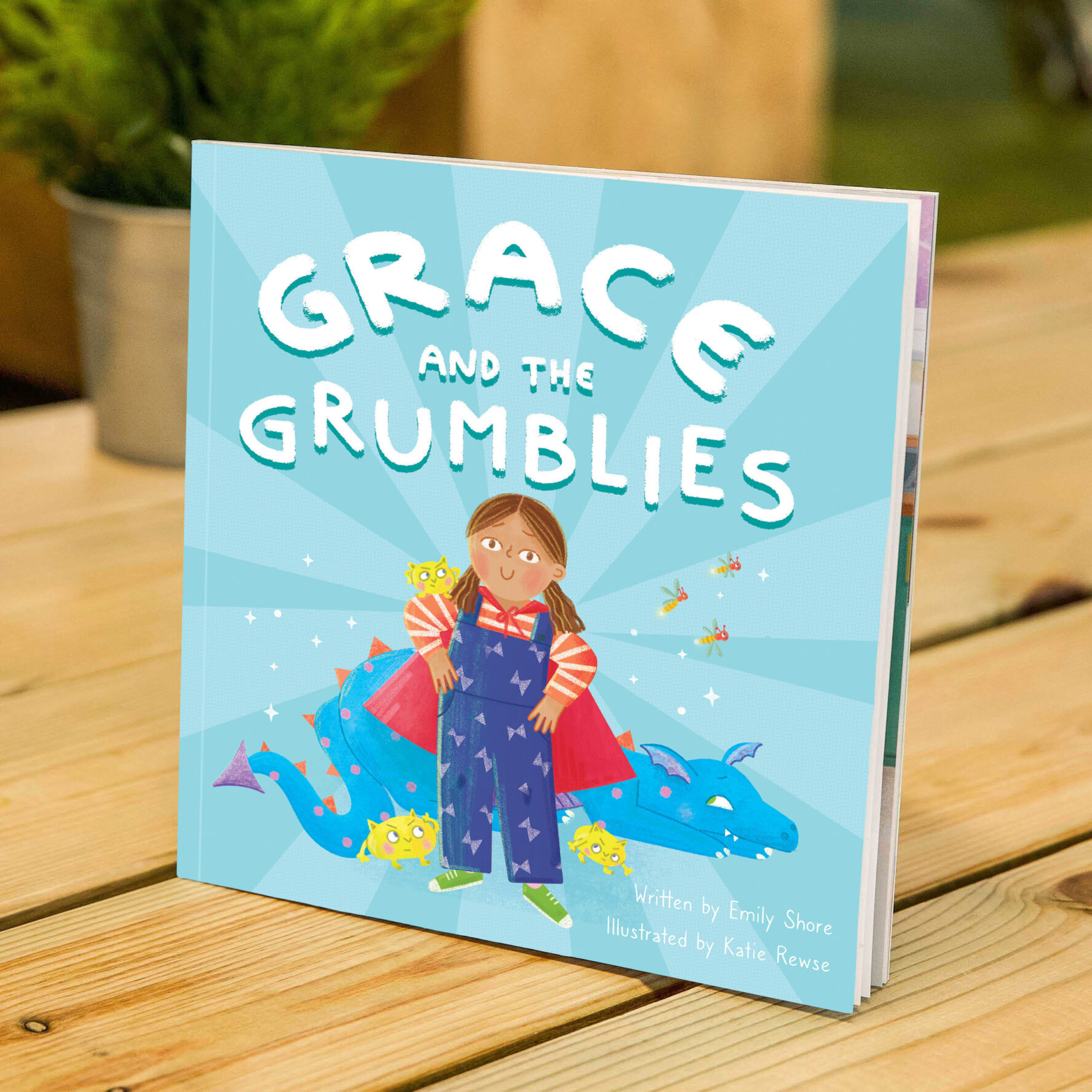 Front cover of 'Grace and the Grumblies' book. A light blue background with a cartoon Grace, a child who is wearing a superhero cape and dungarees. Behind her is a dragon.