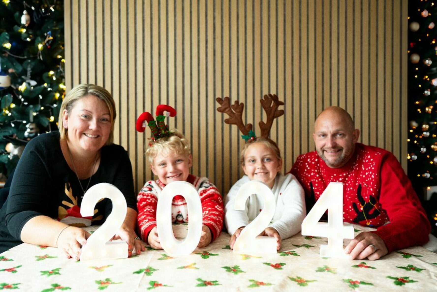 Family of two adults and two children, sat in front of the Christmas tree wearing Christmas jumpers, holding a '2024' sign.