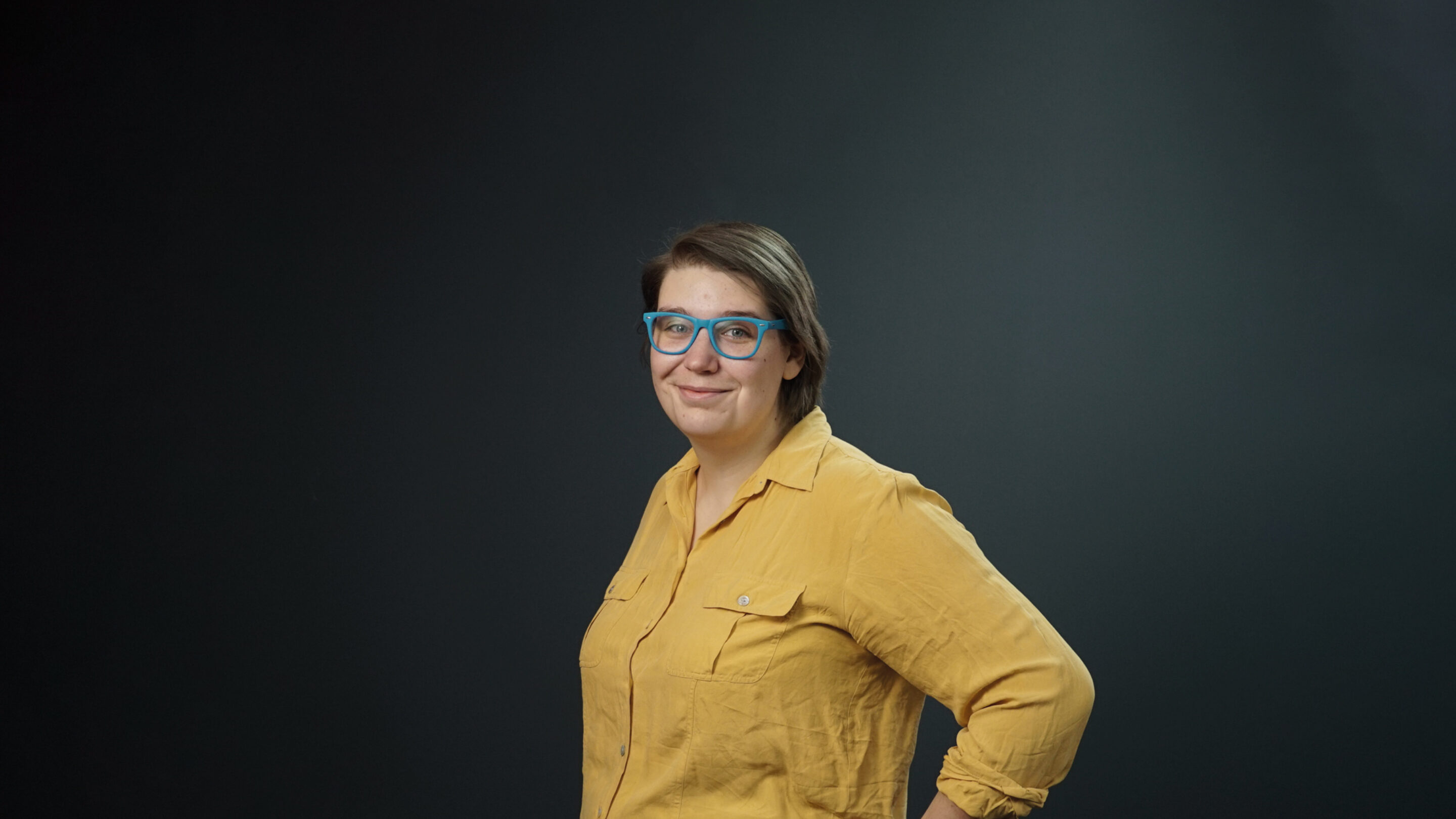 Young woman stood sideways looking towards the camera wearing bright blue glasses.