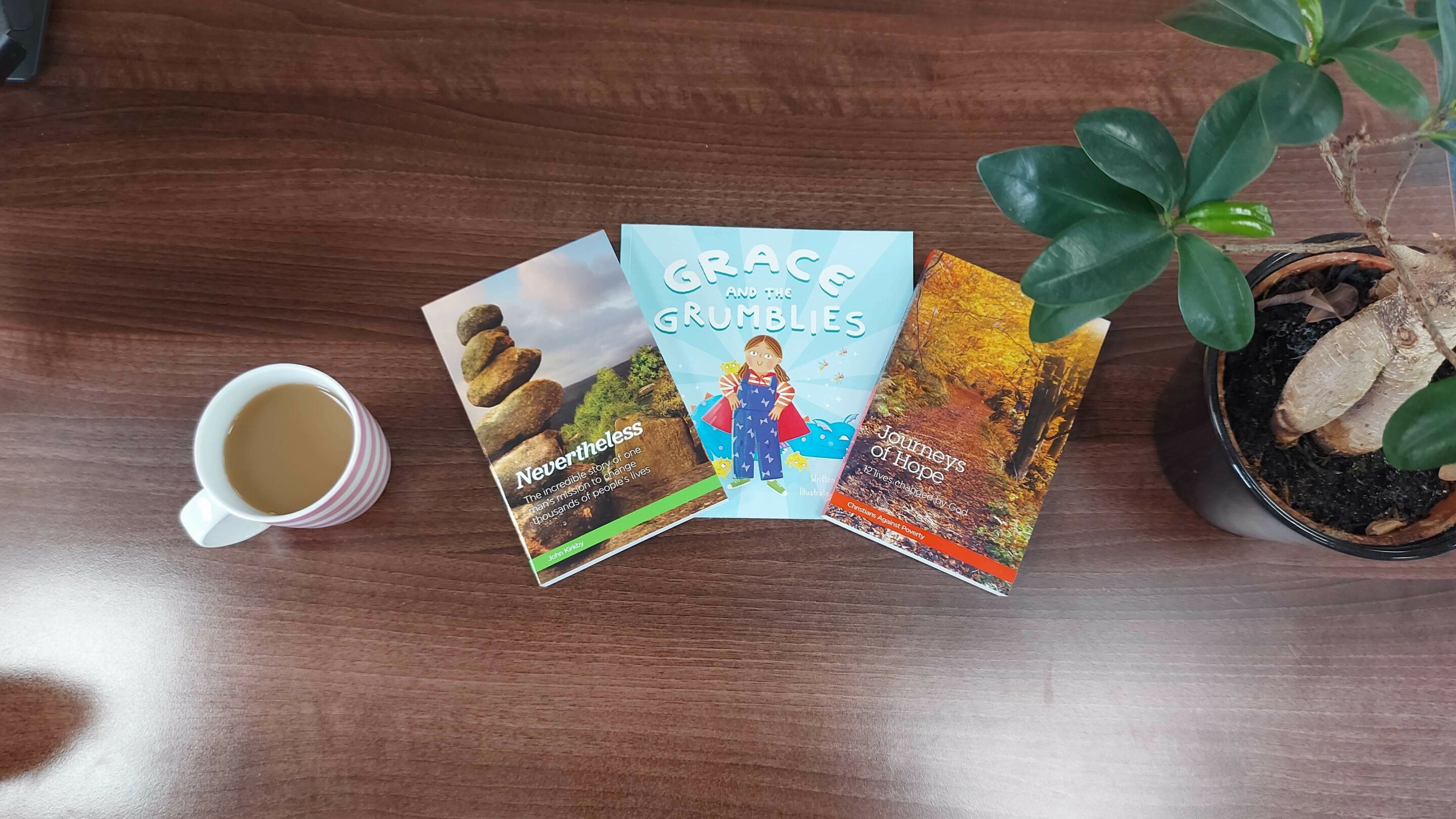 Three books laid out on the table, Nevertheless, Grace and the grumblies and journeys of hope. There is a cup of tea on one side and a plant on the other side.