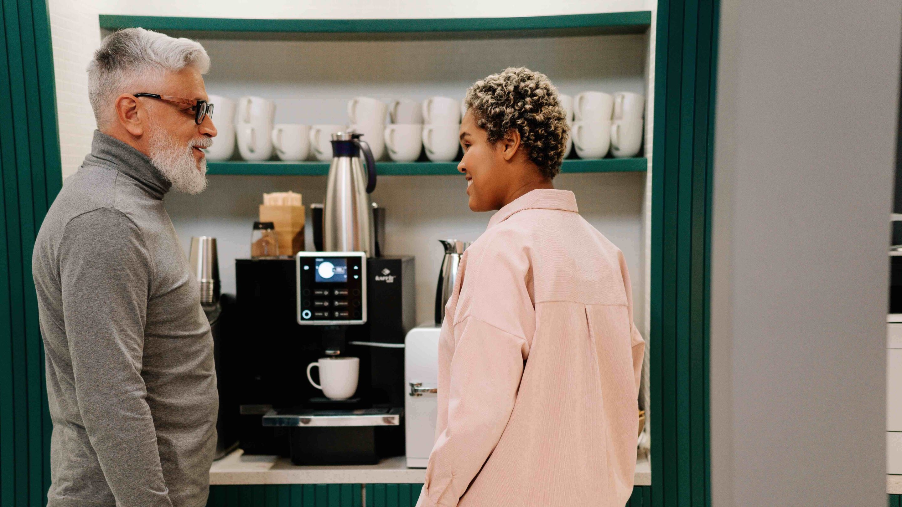 Older male and younger woman stood by a coffee machine chatting while they wait.