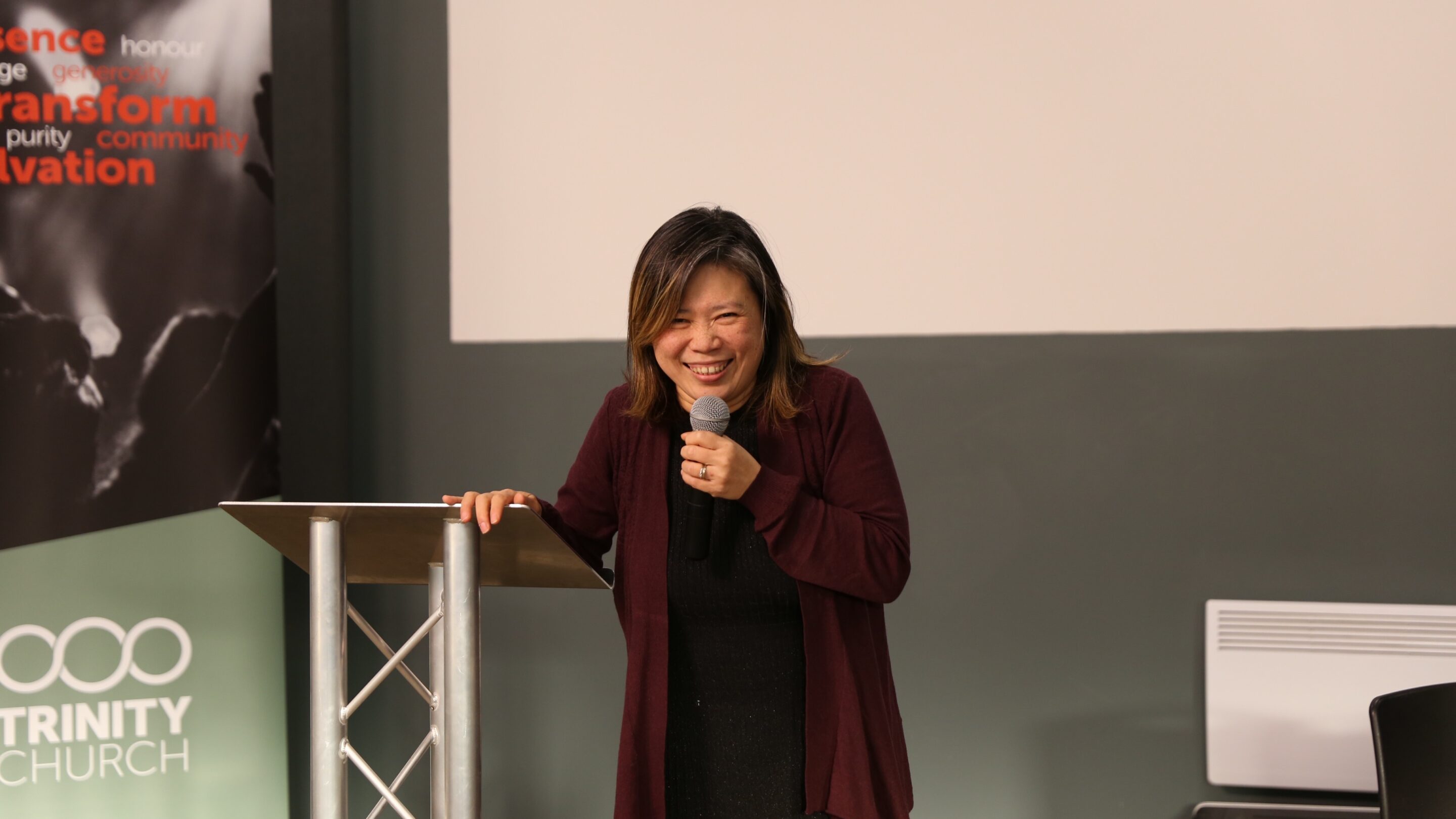 Petite older woman stood with a microphone next to a lectern and smiling.