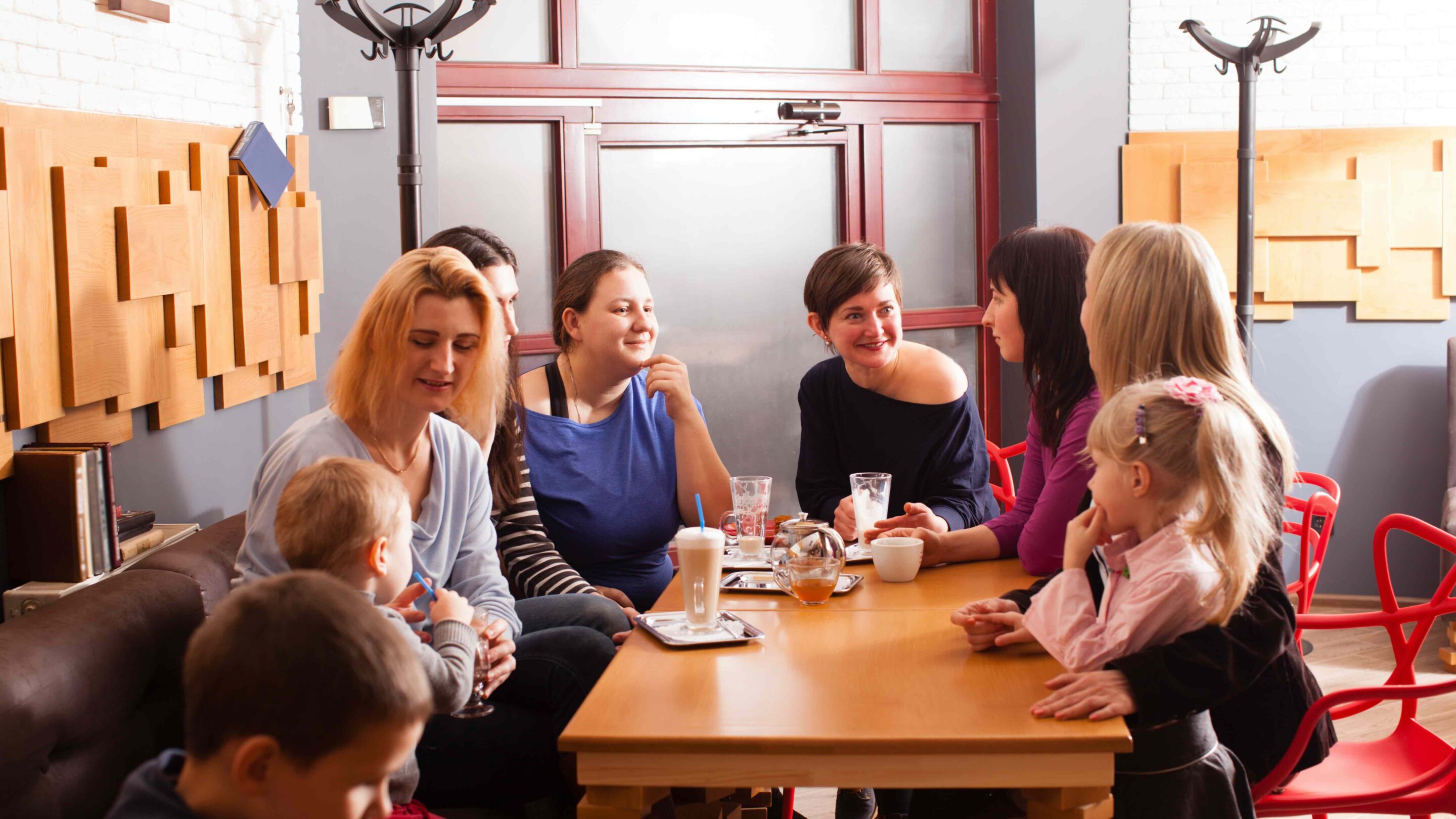 Large group of young women and children sat around a table, talking and smiling with each other.