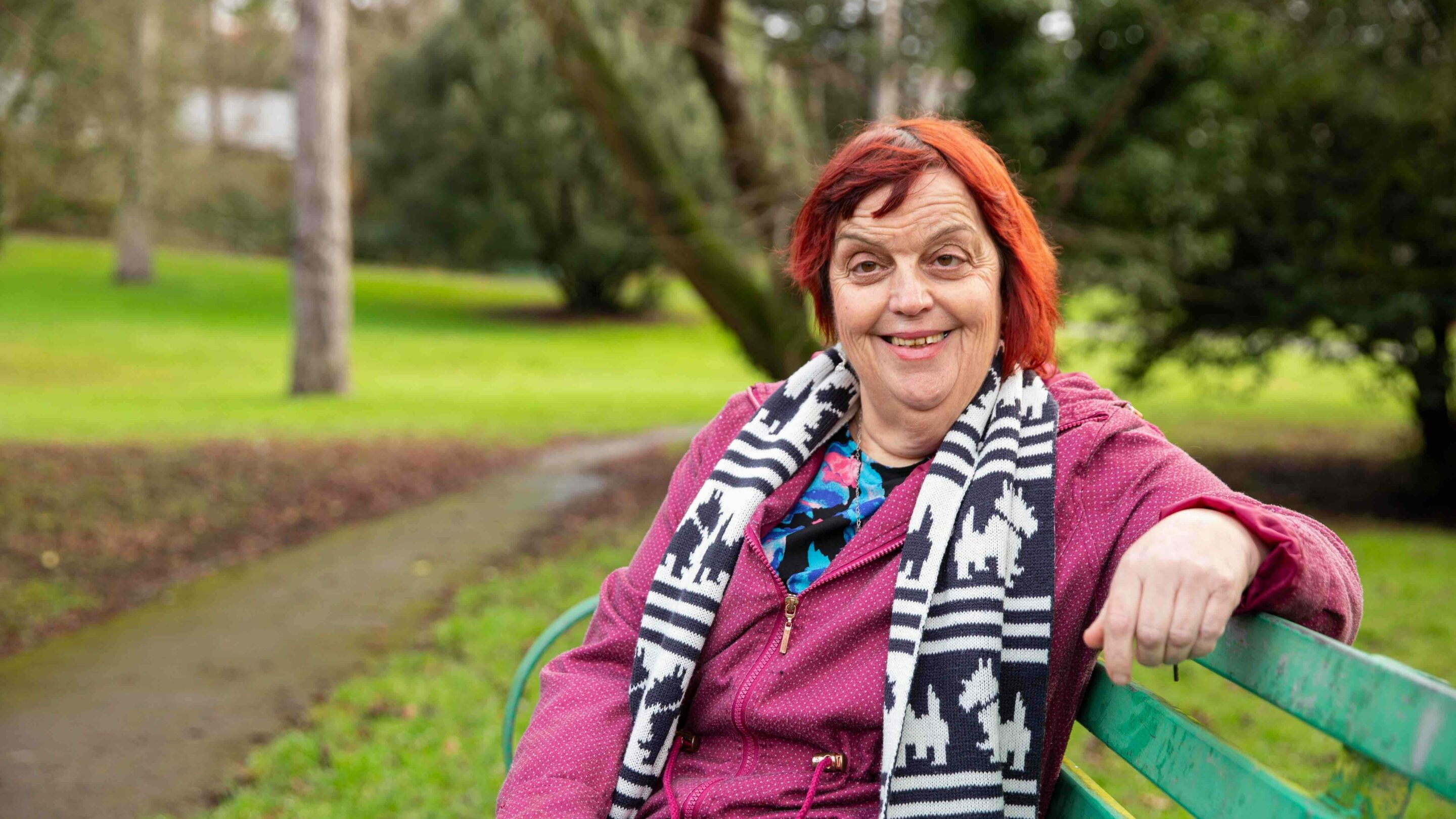Older woman sat on a park bench smiling at the camera.
