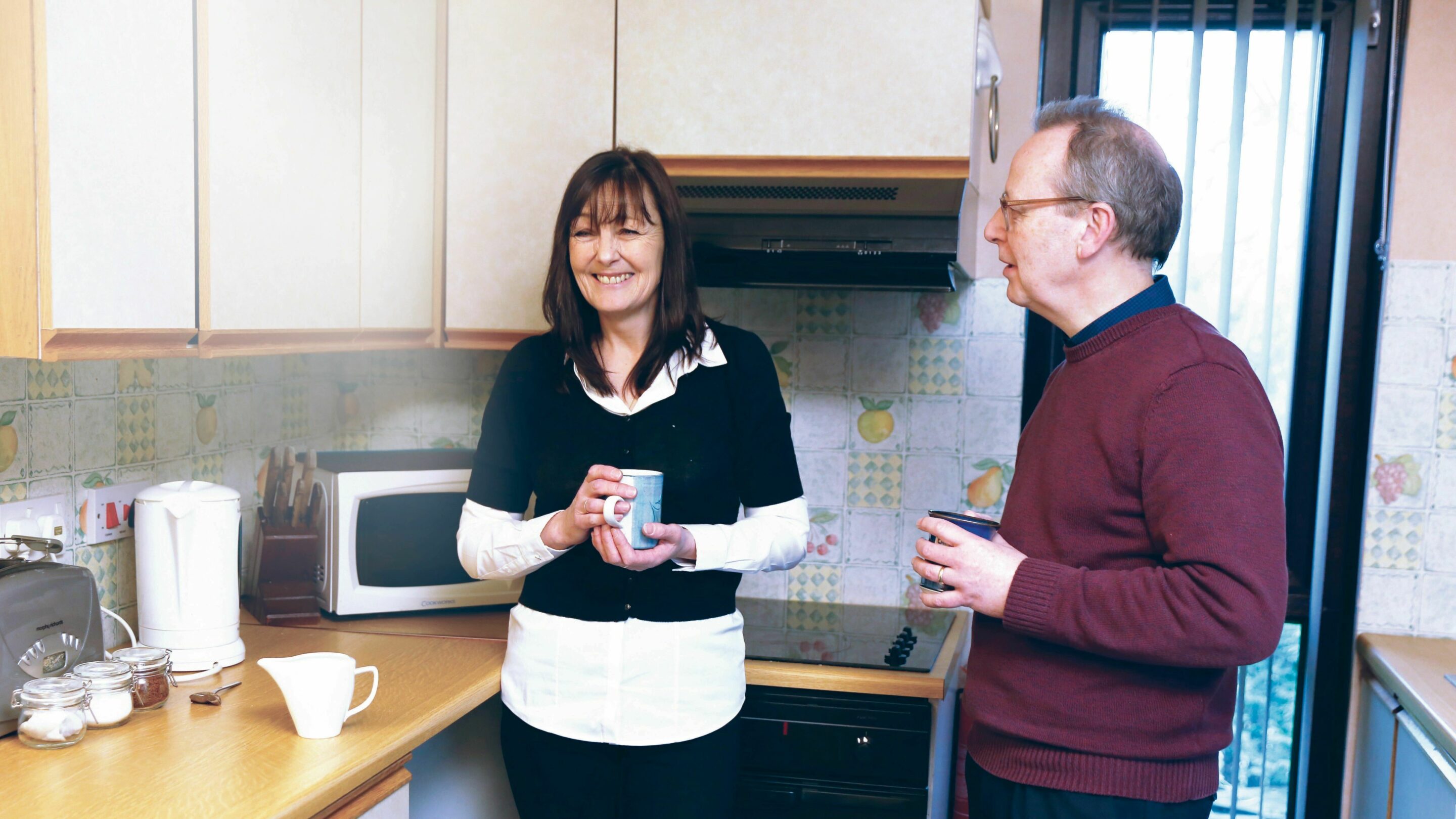 Older male and female stood in the kitchen chatting while holding cups of tea and smiling.