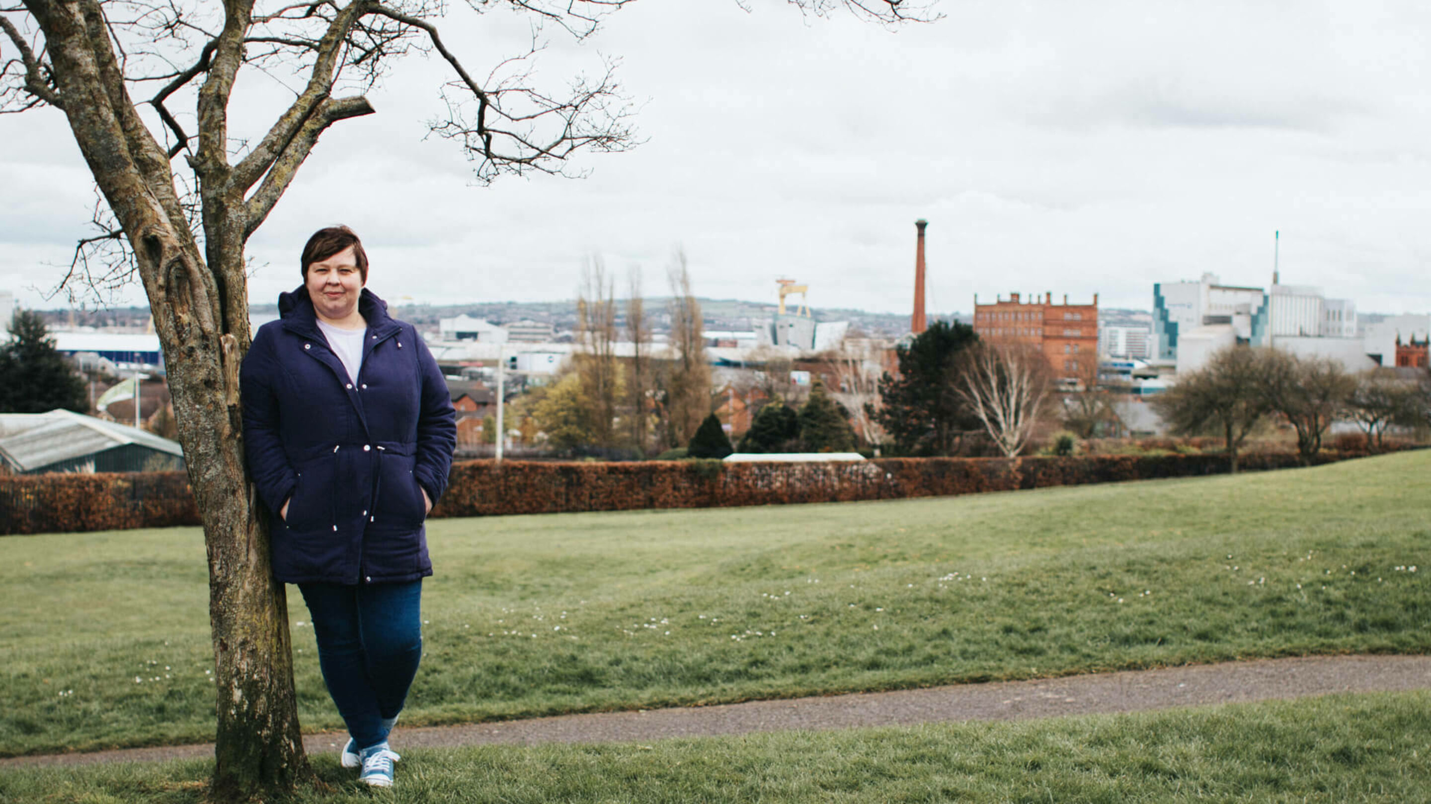 Older woman outside, leaning against a tree with a backdrop of a city in Northern Ireland.