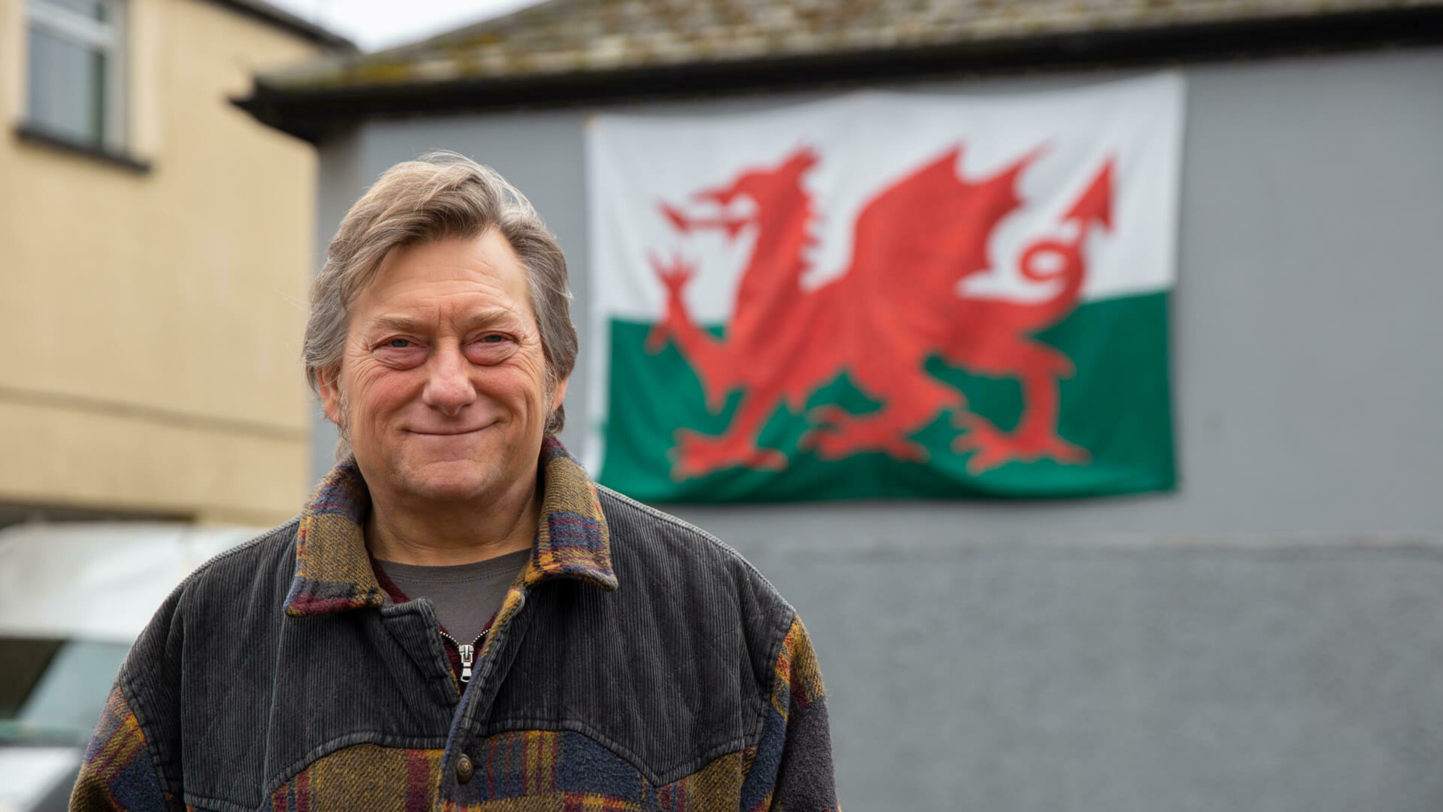 Older male smiling towards the camera with a Welsh flag hung behind him on the side of a building.