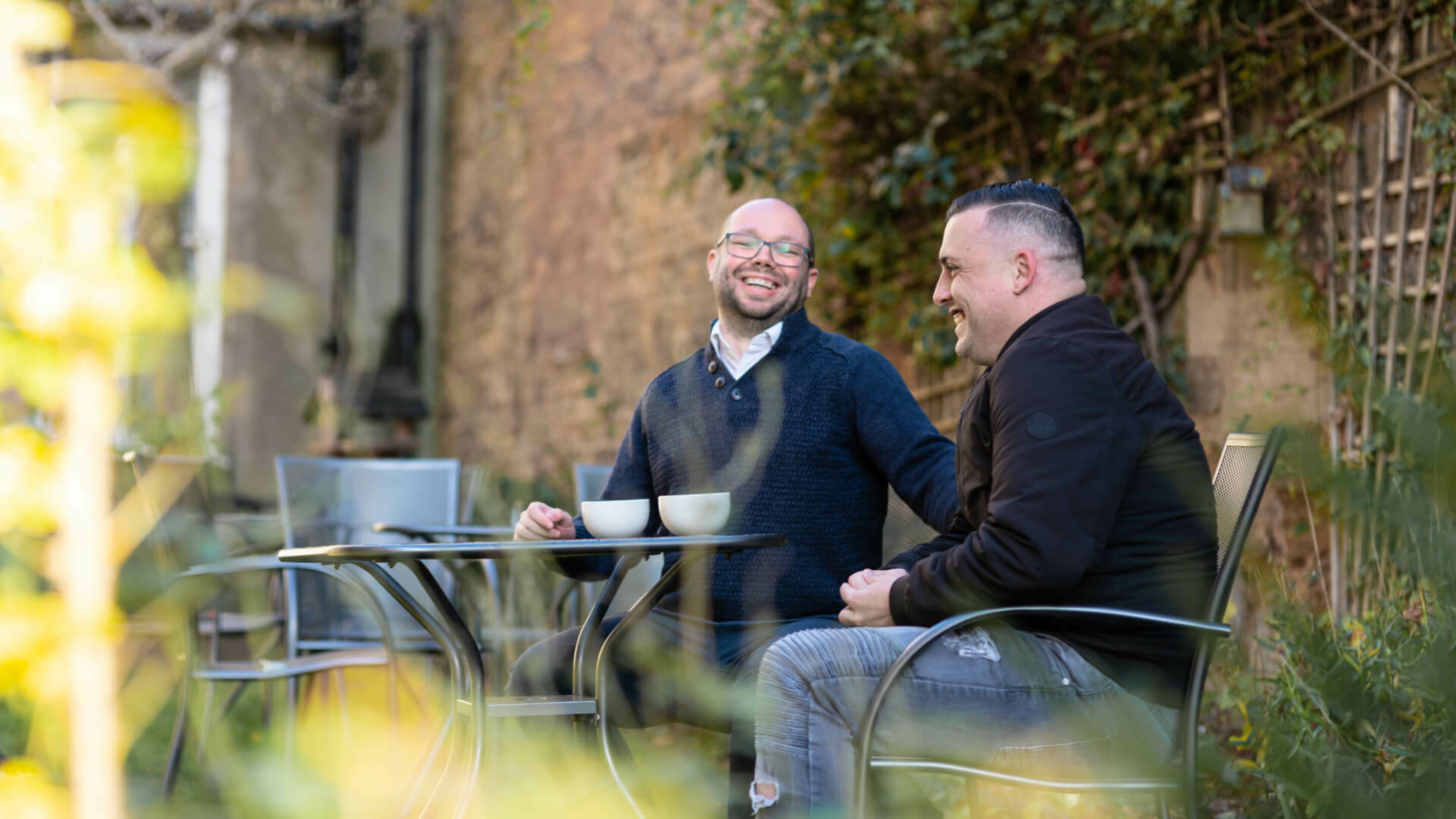 Two young males sat outside drinking hot drinks while laughing and chatting to each other.