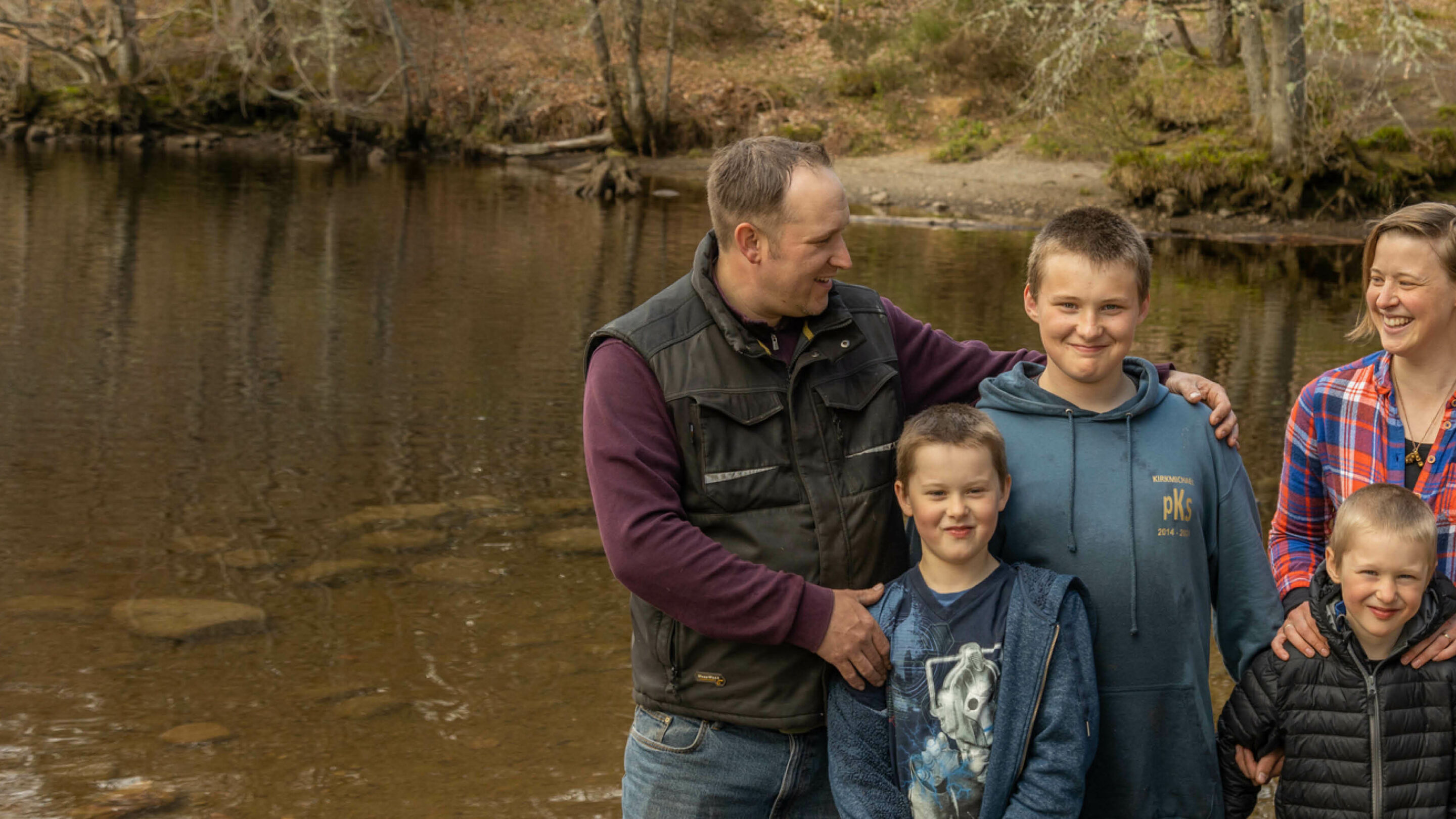 A family of five (mum, dad and three sons) smiling at each other against a background of a river in woodland
