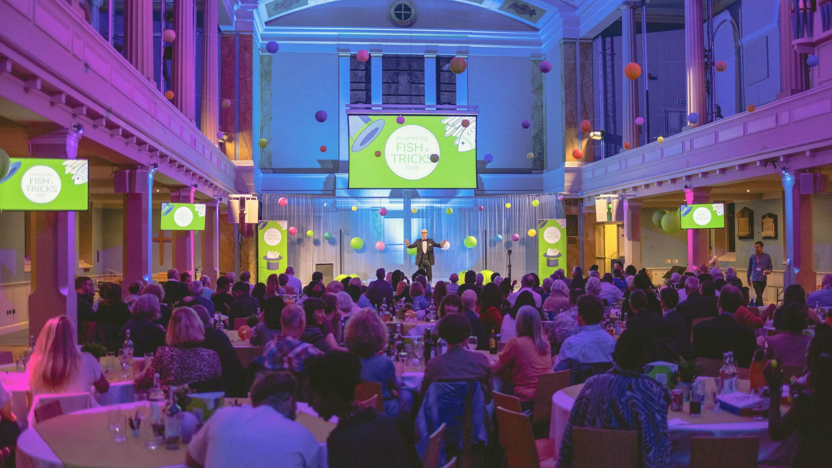 Big conference hall filled with people sat down at tables, purple and blue lights with TV screens presenting 'The great big fish and tricks night'.