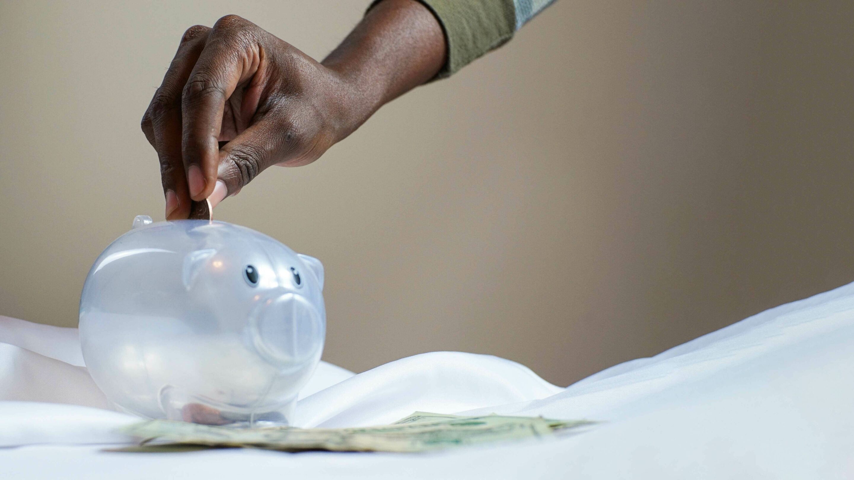Hand holding a coin, placing it into a transparent piggy bank.
