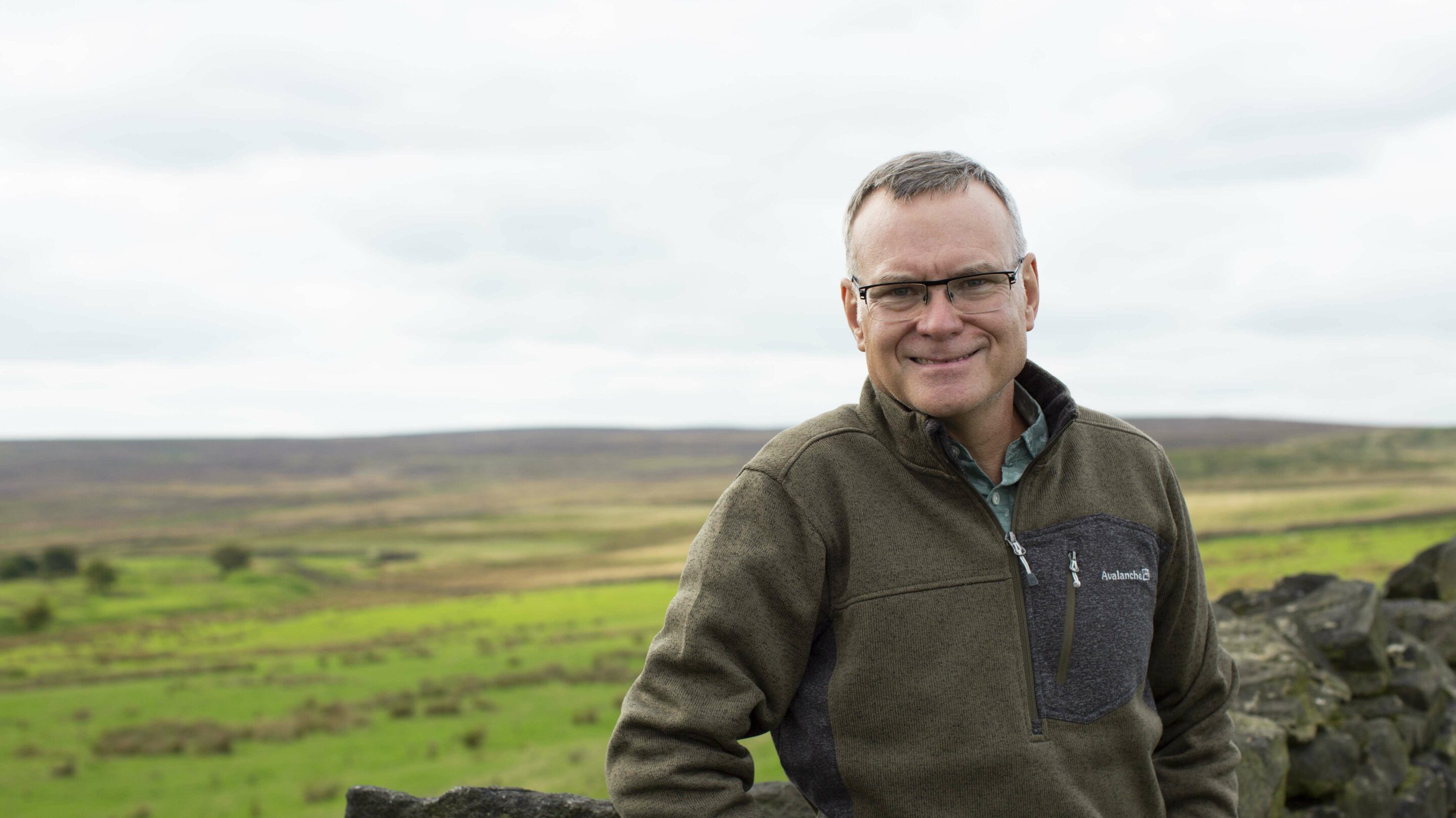 Older male stood outside smiling at the camera, with green fields behind him.