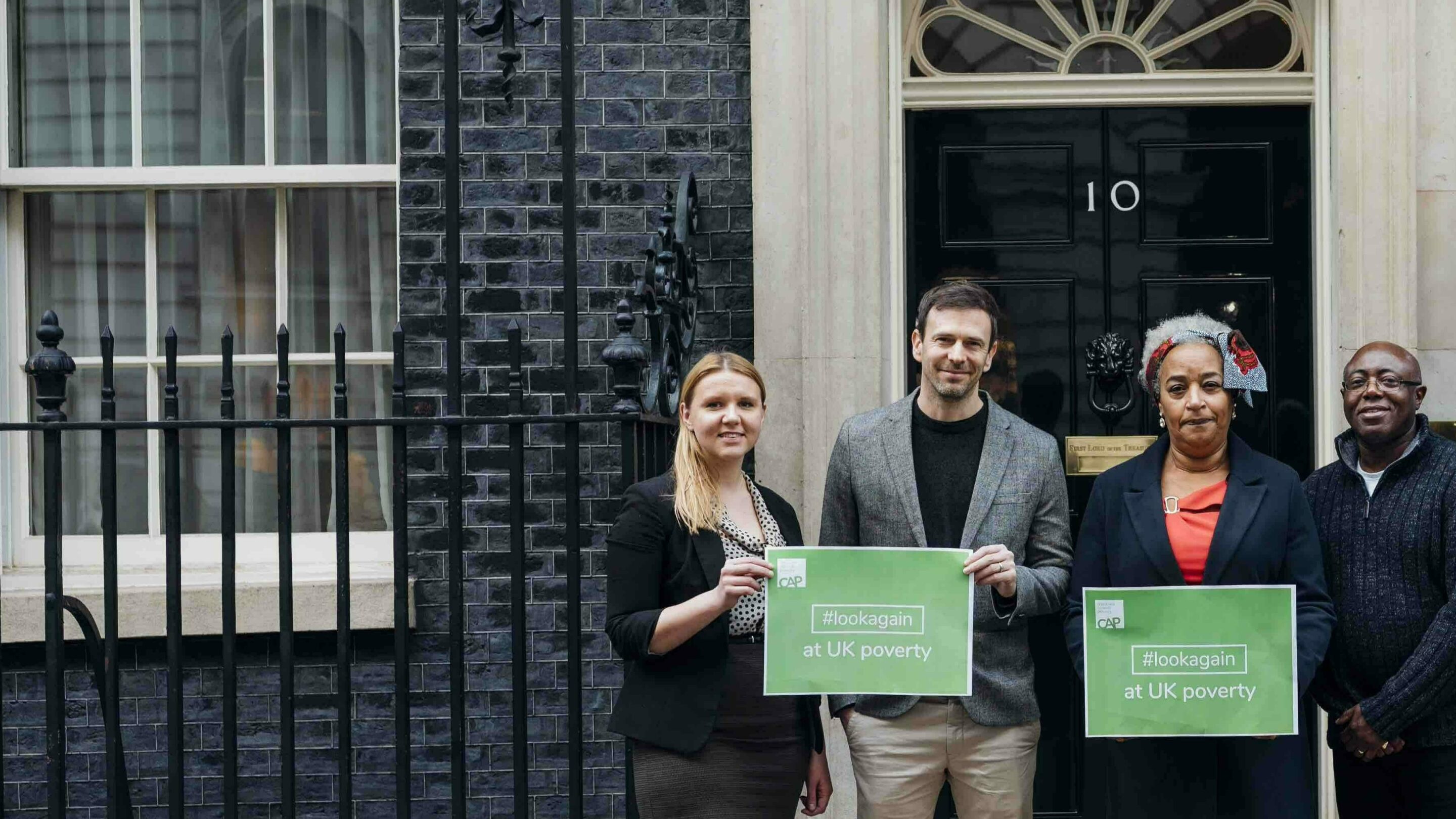Two men and two women stood outside number 10 downing Street holding posters which say, hashtag look again, at UK poverty.