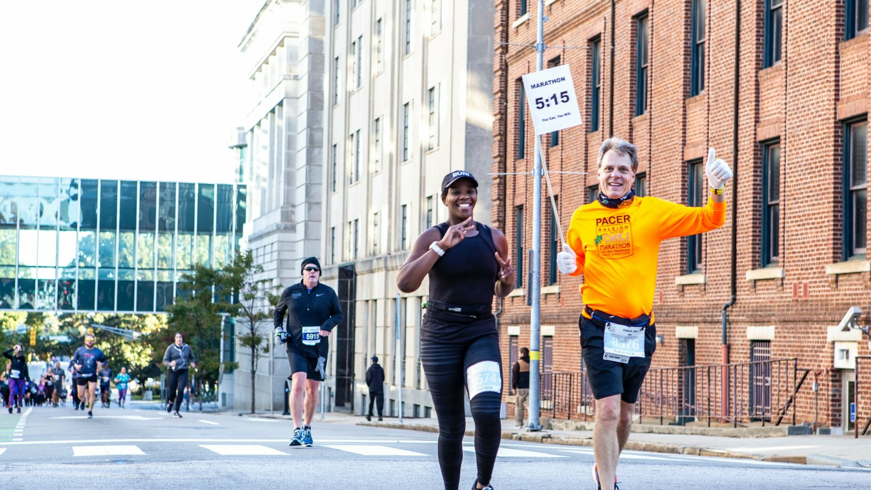 An older male with his thumbs up and younger woman waving as they run in a marathon smiling at the camera.