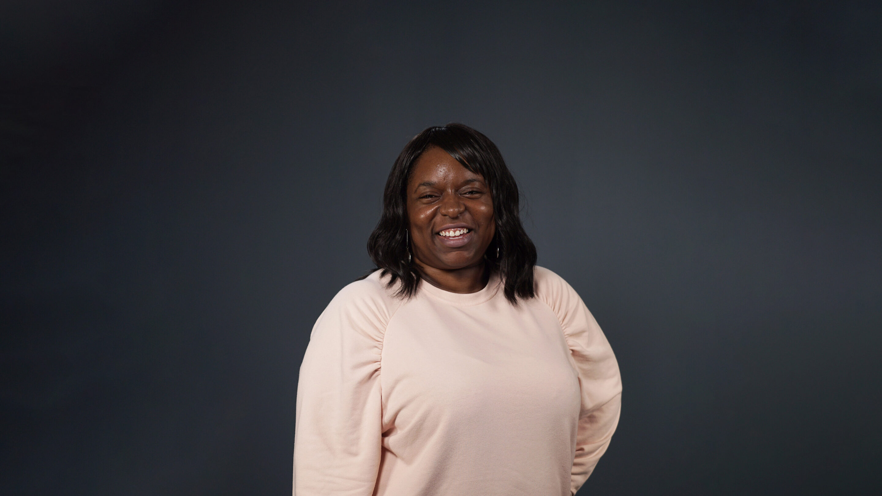 Older woman stood facing forward with her hand on her hip and smiling broadly.