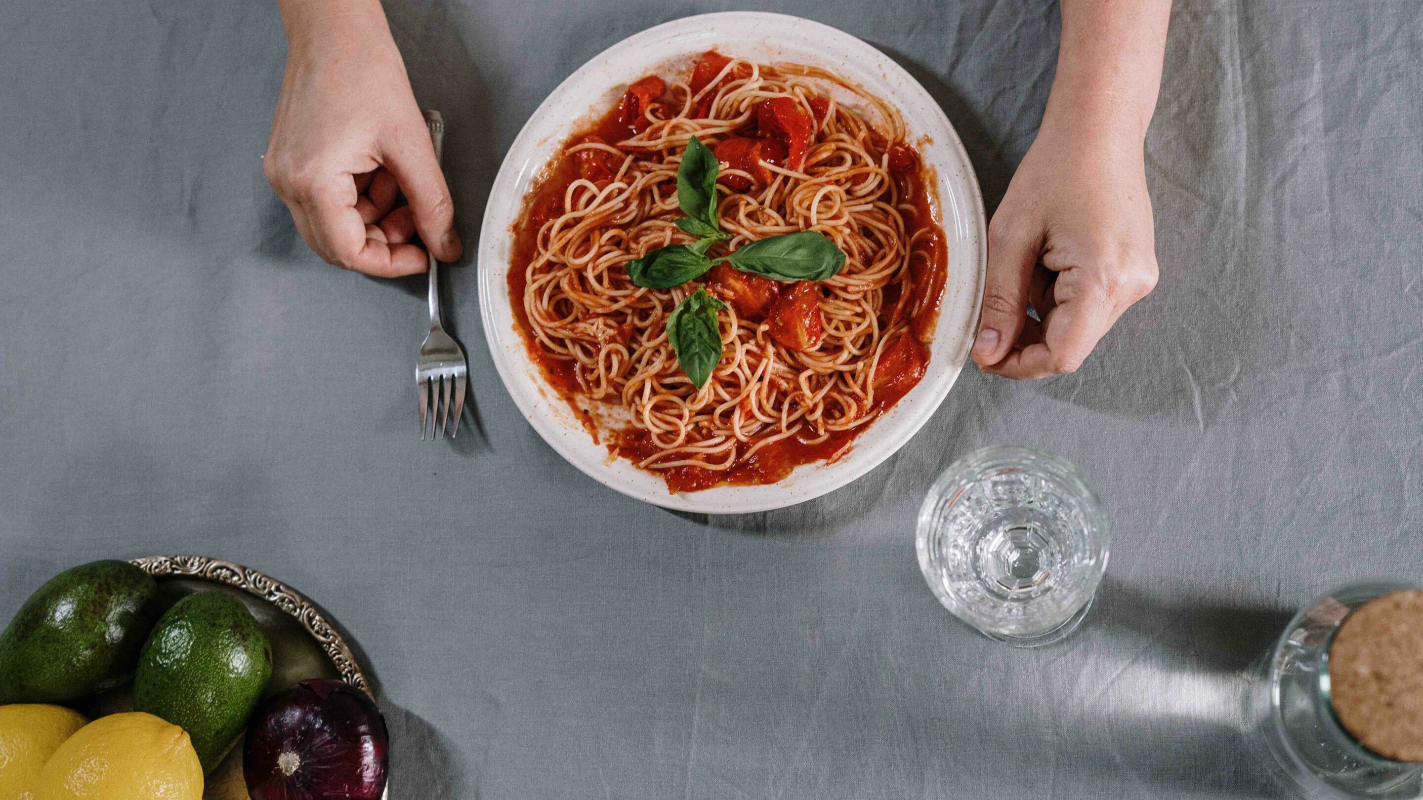 Large bowl of spaghetti bolognese with a glass of water on the side alongside a bowl of fruit in the corner.