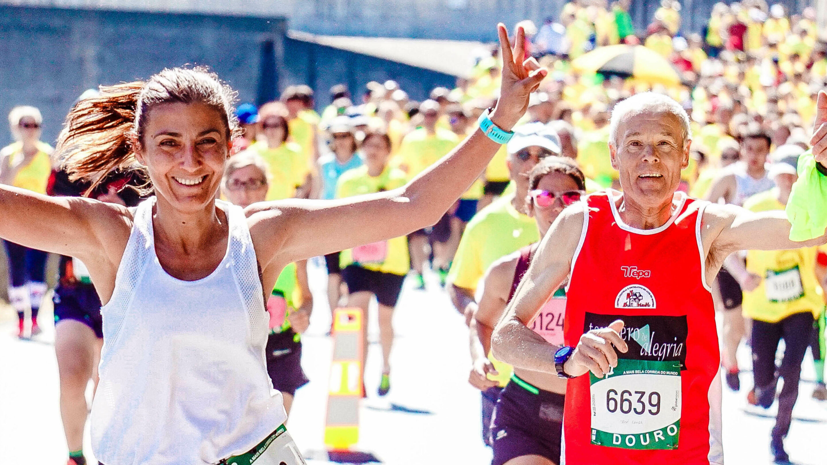 Large group of runners participating in a marathon, one older man has his thumb up, a younger woman has her arms raised in happiness and smiling at the camera.