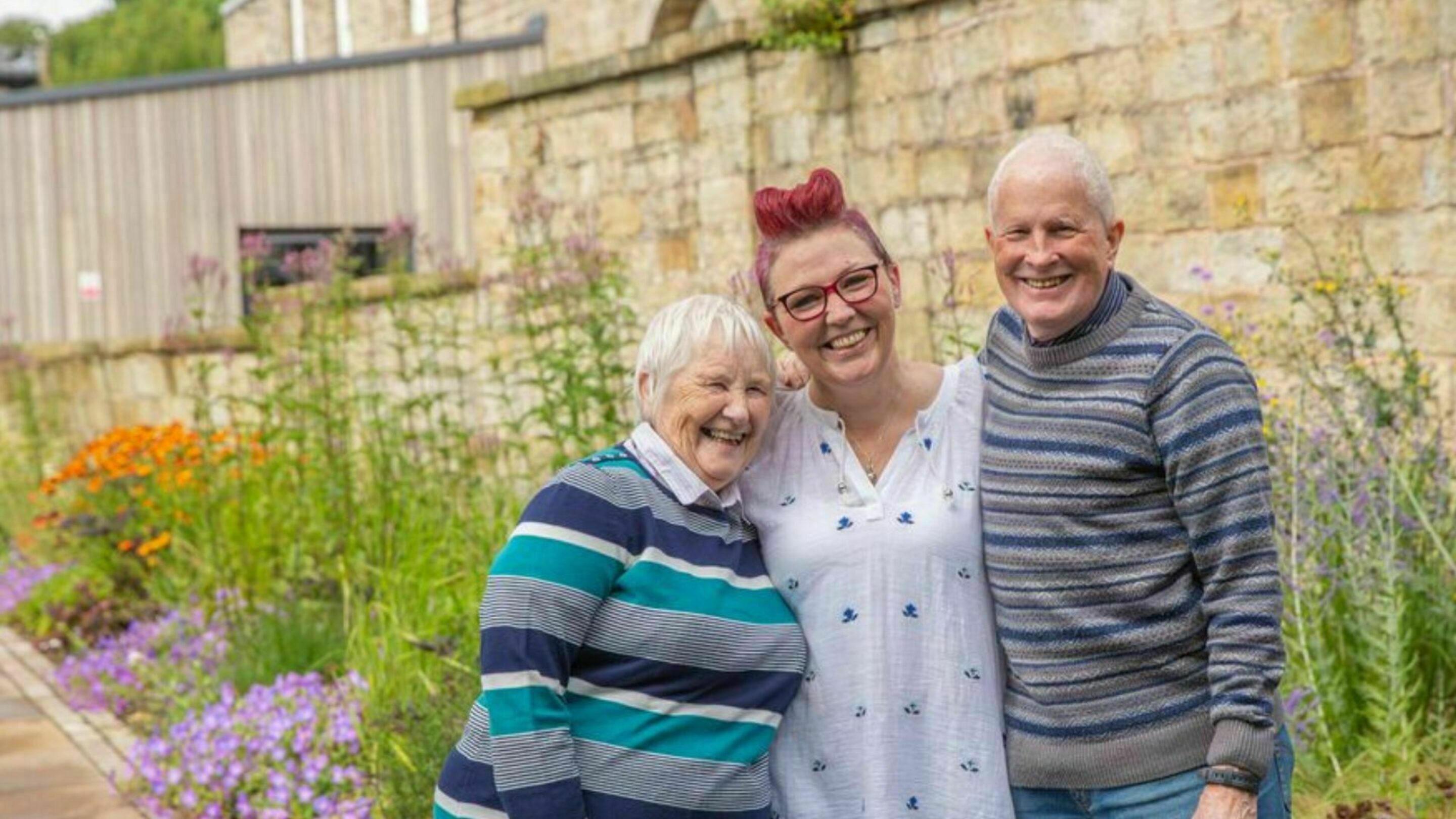 Three people smiling together in a garden. First there's Amanda (centre), former CAP client who became debt free in 2021. She is smiling at the camera. On her left is Mary, and on her right, Malcolm, who were both part of her debt free journey with CAP.