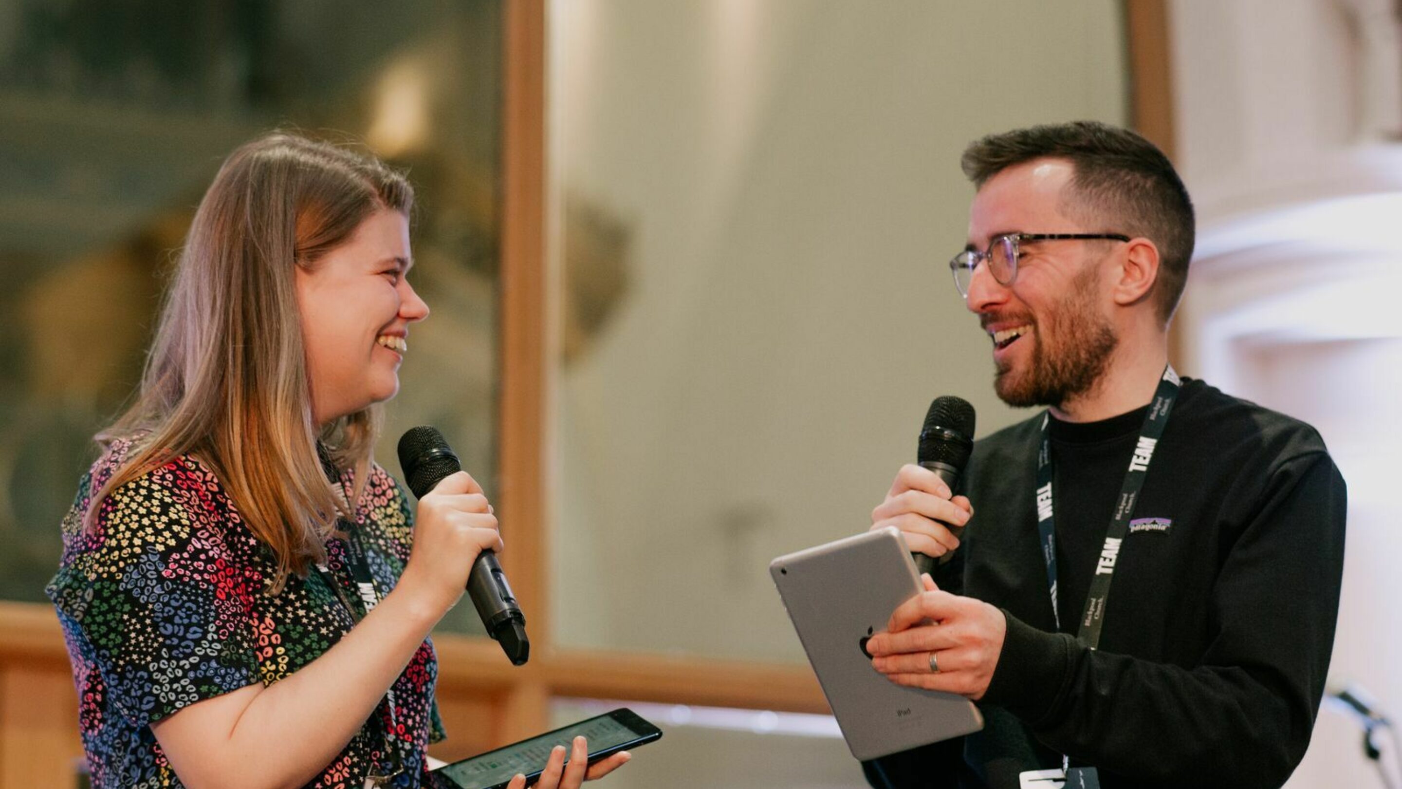 Photo showing two people holding microphones on a stage, looking at each other and smiling