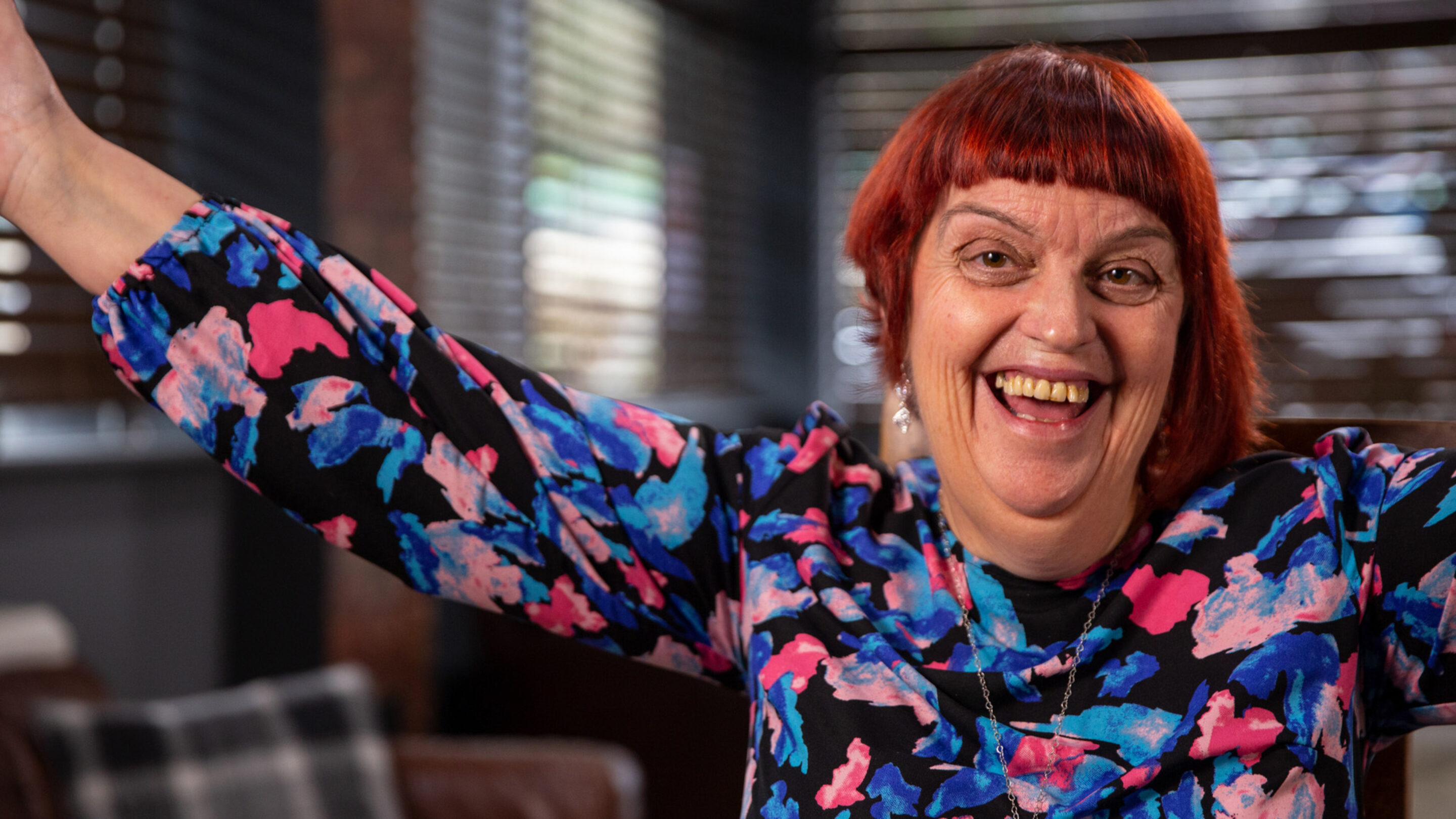 A former CAP client, now debt free. She has bright red hair and colourful top, and is smiling enthusiastically with her hands in the air.