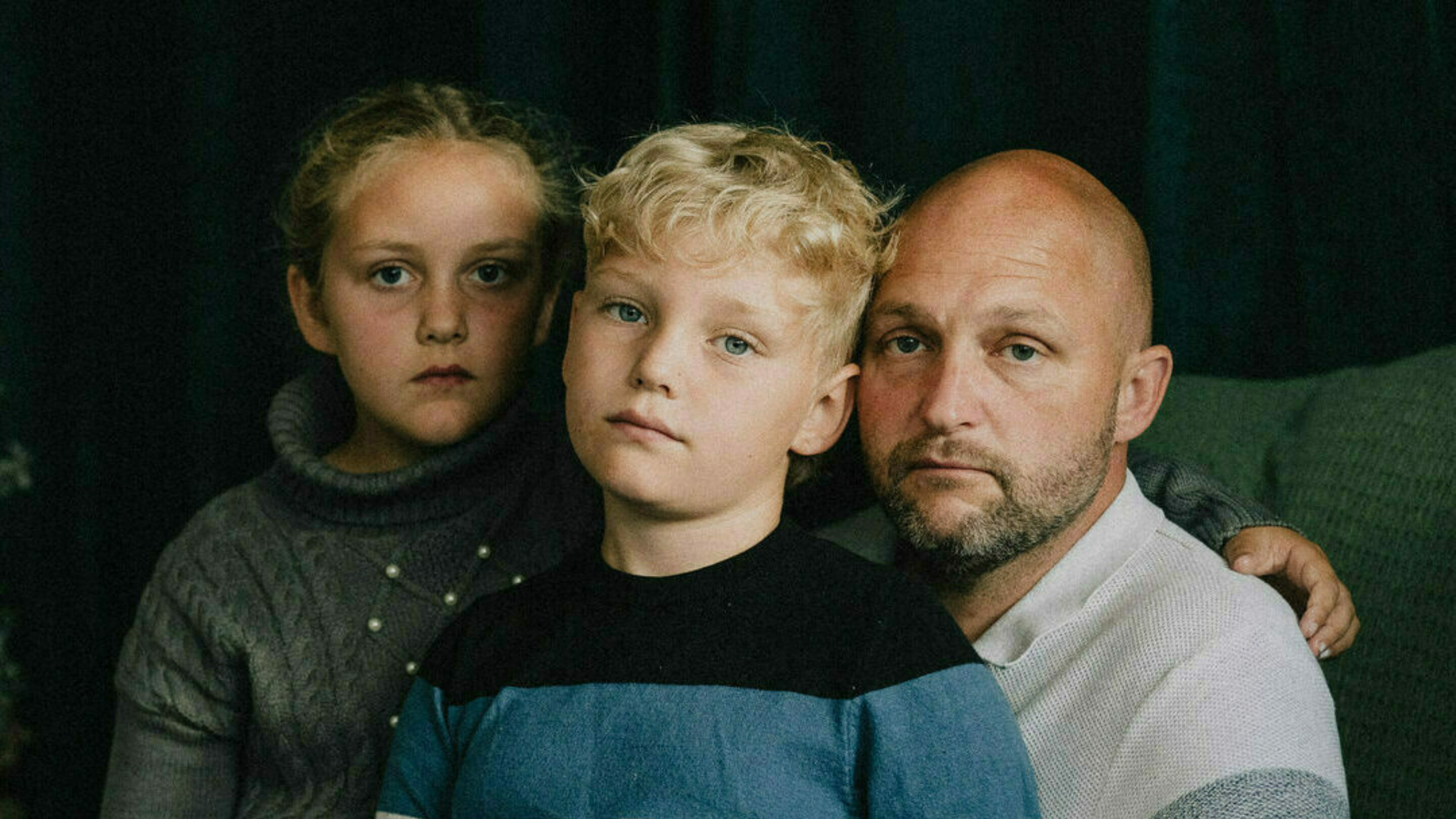 Dad sat with his two young children, all three are staring at the camera with sadness next to a Christmas tree