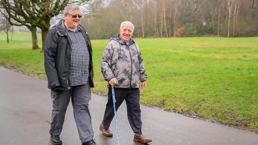 Two older men walking in a park, smiling at each other. One is using a walking stick