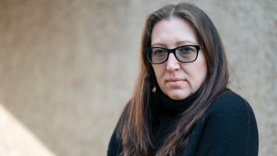 A woman with long brown hair and dark framed glasses looks directly at the camera