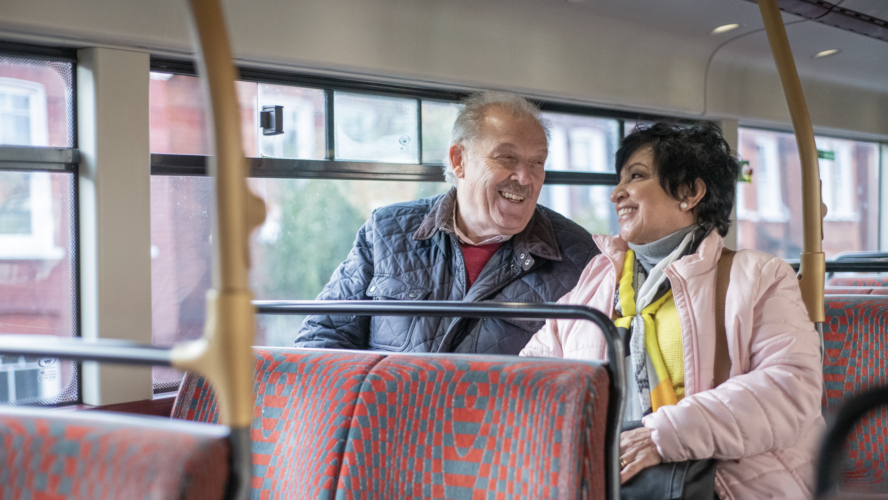 A man and woman sat upstairs on a double decker bus, smiling at each other