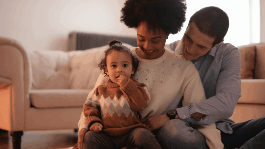 A little girl (aged around 2) facing the camera. She is sitting on her mother's lap, with her father hugging them both from behind.