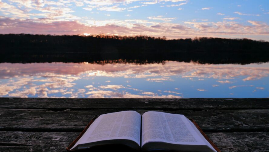 Image of an open Bible on a bench in from of a lake, with the sunset in the distance