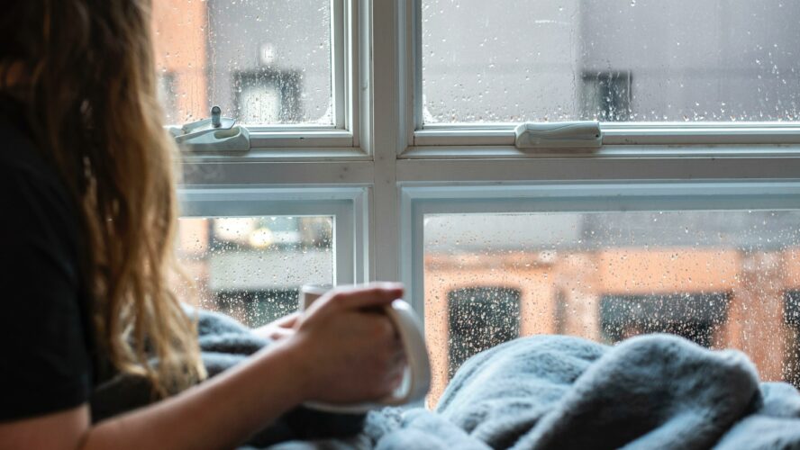 Woman sat at window, in a blanket with hot drink