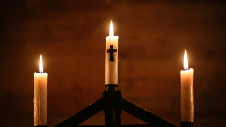 Three pillar candles lit up on a stand. The middle candle has a cross on the front.