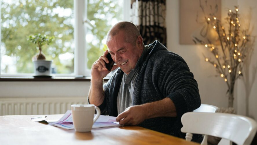 Man with his phone next to his ear and smiling broadly while looking down at his table, holding paperwork.