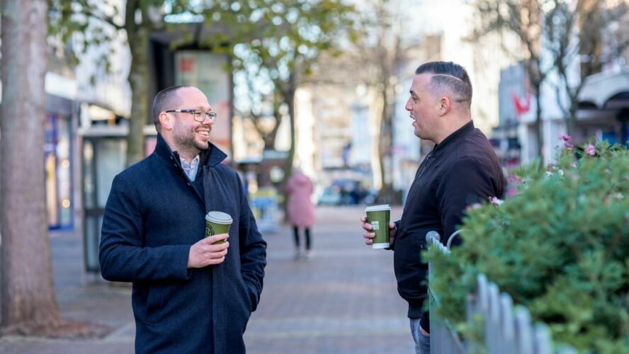 two men drink coffee while chatting on a high street