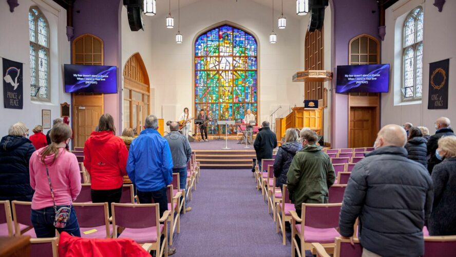 Lowe Church. Taken from the back of the centre aisle, this photo shows the inside of the hall used for church services, with church members stood to worship. They are stood in rows of pink cushioned chairs and straight ahead is a stained-glass window of the cross.