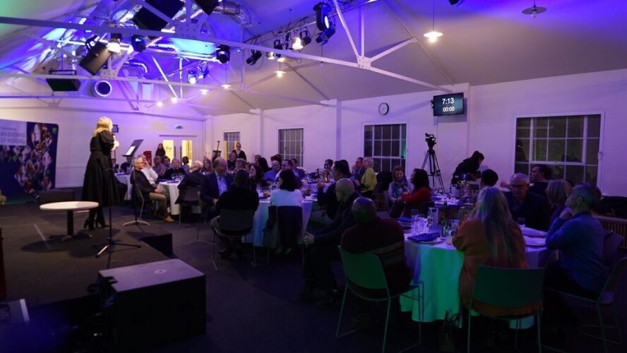A room full of people at an event. Someone talking with a microphone on stage