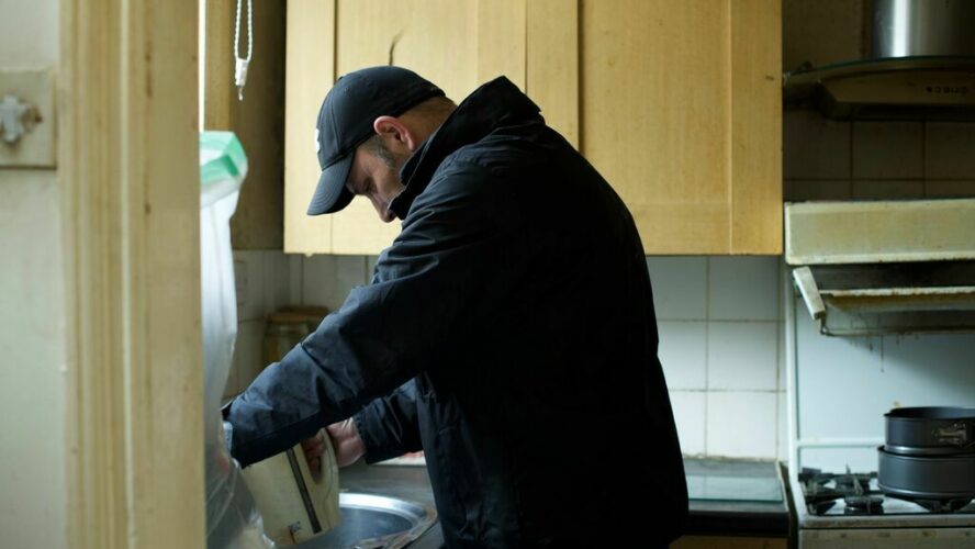 A man wearing a baseball hat filling up a kettle at a sink in a kitchen.