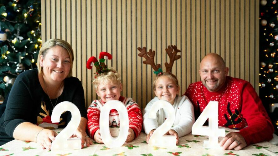 Family of two adults and two children, sat in front of the Christmas tree wearing Christmas jumpers, holding a '2024' sign.