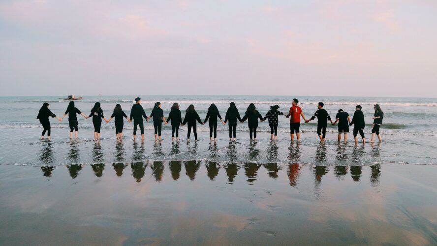 A row of people stand looking out to sea and holding hands