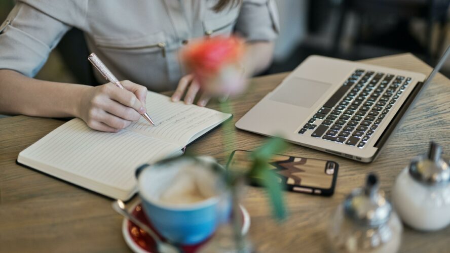 A woman taking notes and drinking coffee watching a webinar on her laptop