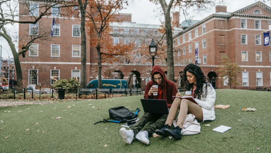 Two students sit on the grass in front of a redbrick building. Both have takeaway coffees and are smiling.