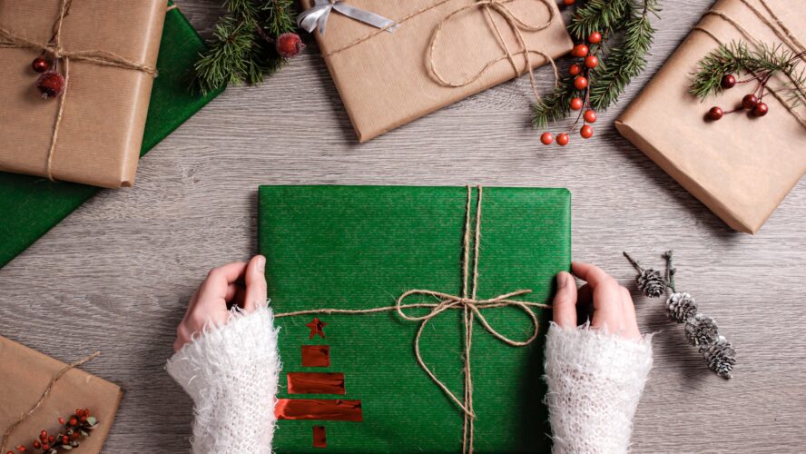 a pair of hands hold a gift wrapped in green paper, surrounded by other parcels and Christmas foliage