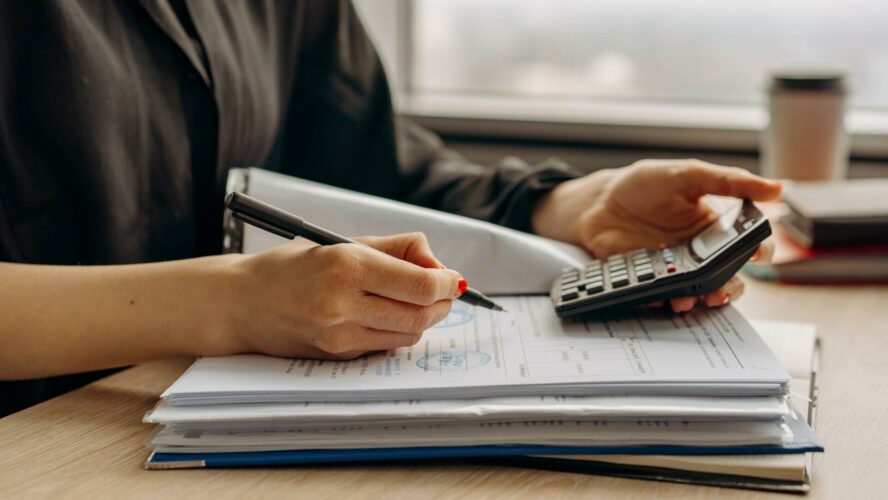 A female's hands holding a pen and calculator on top of a stack of papers