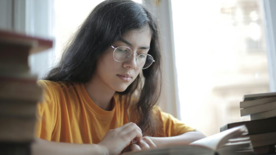 Image of a young person wearing glasses sat down at a table, looking down at a textbook.