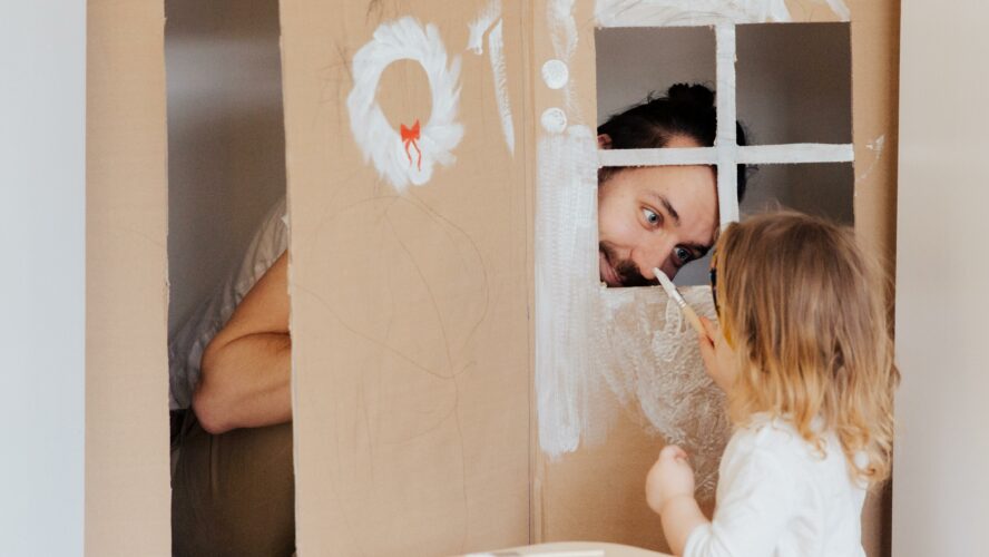 A dad playing in a cardboard house with their child.