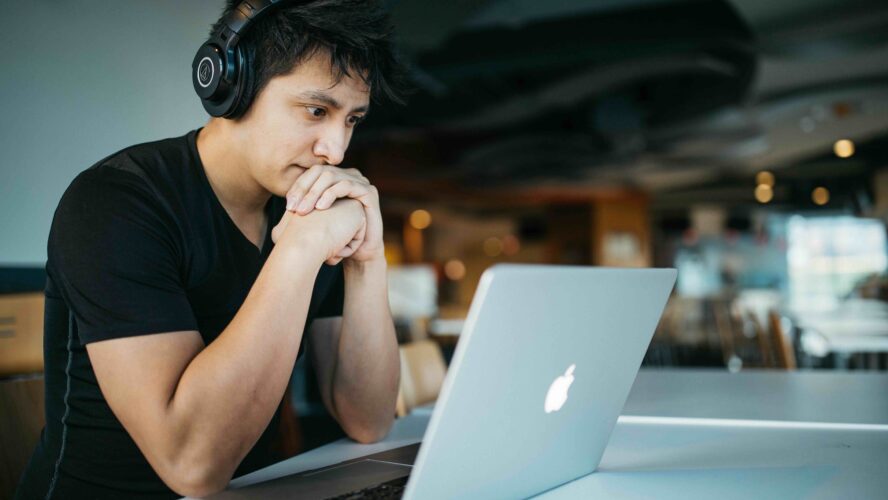 A man sat at a table wearing headphones looking intently at a laptop.